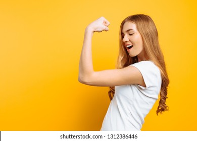 Portrait of happy girl in white t-shirt showing arm muscles on yellow background.