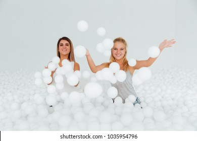 Portrait Happy Girlfriends stand surrounded by white plastic balls in the dry pool. Spend fun time. Adult childhood at Playground with pit-ball. Throw balls in the camera frame.