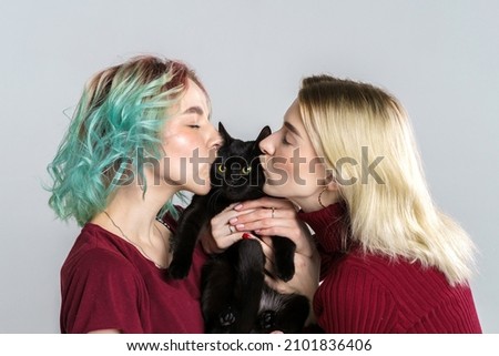Portrait happy girls in casual clothes on light background, holding black cat in their hands and hugging him, smiling and kissing. Cheerful friends hug the cat and rejoice.