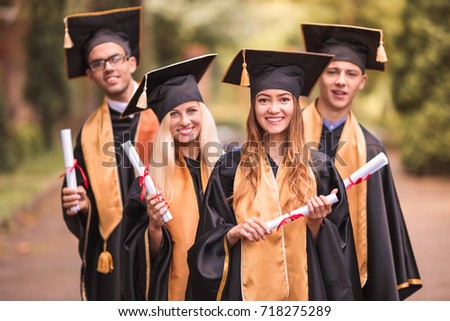 Portrait of happy graduates in mantles who standing in the park and keeping diplomas in hands