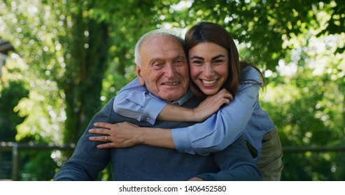 Portrait of happy granddaughter is hugging her grandfather in a wheelchair as a sign of love and respect in a green park on a sunny day.