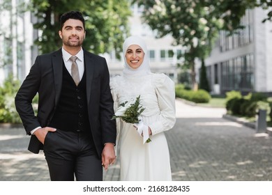 portrait of happy groom holding hands with smiling muslim bride in hijab with wedding bouquet