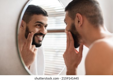 Portrait Of Happy Handsome Arab Man Looking At Reflection In Mirror In Bathroom And Touching Face, Attractive Masculine Middle Eastern Guy Putting Cream On His Cheek. Male Facial Care Concept - Shutterstock ID 2159141445