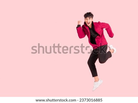 Portrait of happy handsome Asian man in a pink jacket excited and celebrating by jumping up in the air with winner gesture isolated on pink background. The young male shows his confidence by jumping.