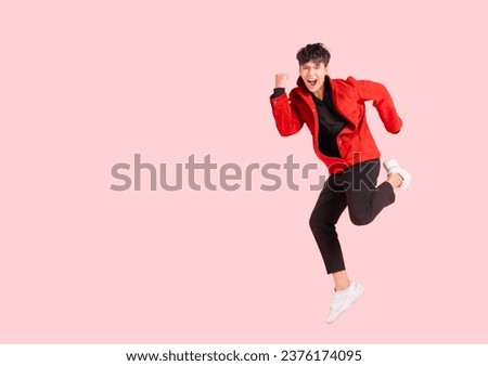 Portrait of happy handsome Asian man in a red jacket excited and celebrating by jumping up in the air with winner gesture isolated on pink background. The young male shows his confidence by jumping.