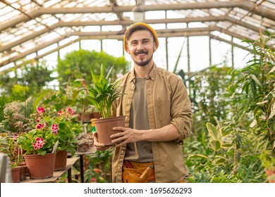 Portrait Of Happy Handsome Guy In Hipster Cap Holding Potted Plant While Choosing Plant In Nursery House