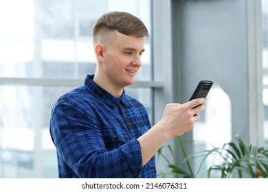 Portrait Of Happy Handsome Guy, Young European Blonde Man In Shirt Is Using His Cell Mobile Phone, Looking At Screen Of Smartphone, Reading Or Typing Message And Smiling In Office Building Indoors.