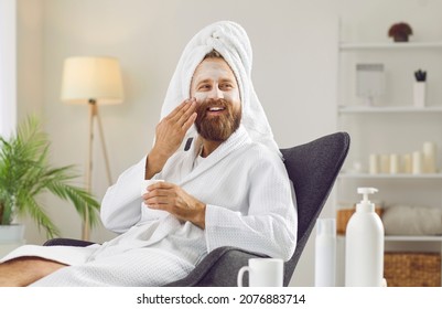 Portrait Of Happy Handsome Unshaven Ginger Man In Fresh Clean Bathrobe And Towel Turban Enjoying Spa And Self Care Day At Home, Relaxing In Chair, Drinking Detox Tea And Applying Beauty Clay Face Mask