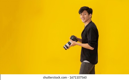 Portrait Of Happy Handsome Young Asian Male Professional Photographer Holding A Professional Digital Camera Isolated Over  Yellow Color Background.