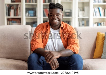 Portrait of happy handsome young black man in casual outfit with braids sitting on couch at home, smiling at camera, living room interior, copy space. Millennials lifestyles concept