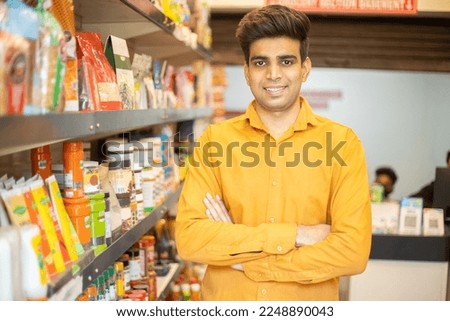 Portrait of happy handsome young Indian man wearing orange shirt standing cross arm at grocery shop or supermarket, Closeup. Selective Focus.