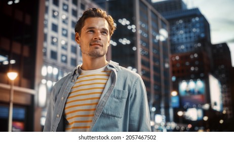 Portrait Of A Happy Handsome Young Man In Casual Clothes Standing On The Street At Sunset. Stylish Male Model In Big City Living The Urban Lifestyle. Background With Office Buildings And Billboards.