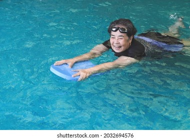 Portrait Of Happy And Healthy Asian Senior Woman Swimming With Kickboard In A Swimming Pool. Smiling Old Woman Swimming With Inflatable Board In Swimming Pool.  Elderly Active Lifestyle.