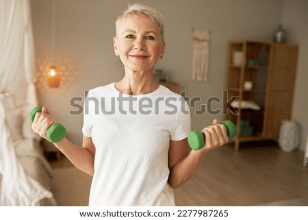 Portrait of happy healthy athletic elderly woman in white mockup t-shirt with copy space for your advertising content exercising at home with green dumbell, training hand and back muscles