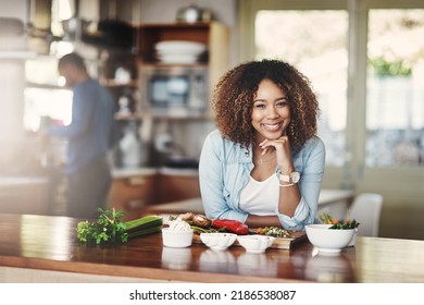 Portrait Of A Happy, Healthy And Carefree Young Woman Preparing A Healthy Meal At Home With Her Husband In The Background. Black Wife Making An Organic Vegetarian Salad For Lunch In A Kitchen