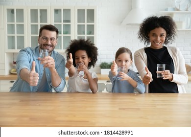 Portrait Of Happy Healthy Mixed Race Family Holding Glasses Of Pure Cool Water, Showing Thumbs Up Gesture. Positive Smiling Emotional Multiracial Couple With Children Recommending Healthcare Habit.