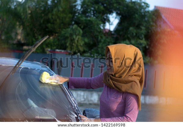 portrait of happy  hijab woman cleaning and washing\
car windshield with water yellow sponge and soap at outdoors area\
on a sunny day