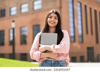 Portrait of happy hispanic lady student posing with laptop in hands outdoors, looking and smiling at camera, having break after classes in university campus - Powered by Shutterstock