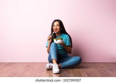 Portrait of a happy Indian asian pretty young woman eating fresh salad from a bowl while sitting isolated against pink wall on wooden floor