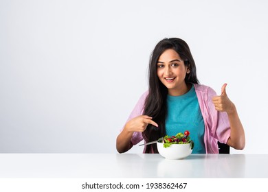 Portrait of a happy Indian asian pretty young woman eating fresh salad from a bowl while sitting isolated at table or desk against white background