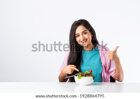 Portrait of a happy Indian asian pretty young woman eating fresh salad from a bowl while sitting isolated at table or desk against white background