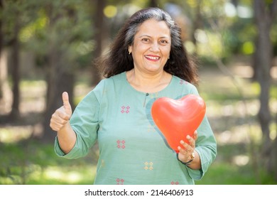 Portrait Of Happy Indian Asian Senior Woman Holding Red Heart Balloon At Park Outdoor Doing Thumbs Up, Good Health Good Life, Elderly An Old People Health Care Concept,