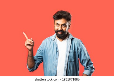 Portrait of a happy Indian bearded young man pointing fingers away at copy space isolated over red background - Powered by Shutterstock