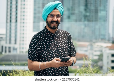 Portrait Of Happy Indian Businessman Using Mobile Phone Outdoors. 
Cheerful Smiling Business Man With Turban Typing Text Message On His Phone And Looking At Camera While Standing In The City.