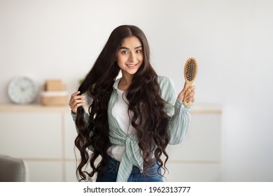 Portrait Of Happy Indian Lady Brushing Her Long Dark Hair With Wooden Brush At Home. Lovely Young Woman Taking Care Of Her Beautiful Locks. Beauty, Wellness And Hairdressing Concept