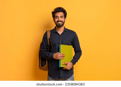 Portrait of happy indian male student with backpack and books posing over yellow studio background, handsome eastern guy holding workbooks and smiling at camera, enjoying modern education, copy space