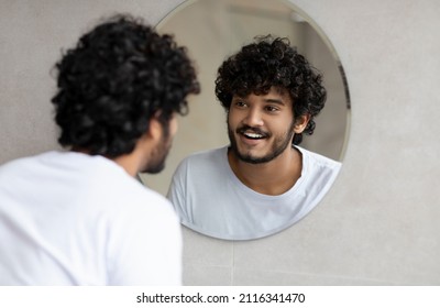 Portrait Of Happy Indian Man Looking At His Reflection In Round Mirror On The Wall, Young Guy Standing In Modern Bathroom At Home, Free Space