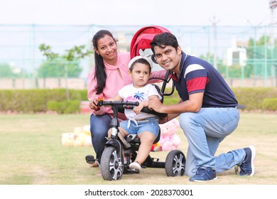 Portrait Of Happy Indian Parents Help Their Cute Little Adorable Daughter Learn Tricycle Sitting In Garden Or Park. Smiling Father Mother Teach Small Girl Child Kids Bike, Enjoy Family Time Together