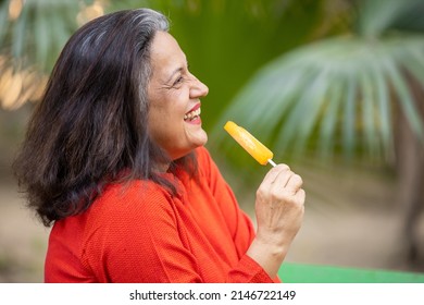 Portrait Of Happy Indian Senior Woman Eating Ice Lolly Or Ice Cream In A Park Outdoor, Old Mature Lady Enjoy Retirement Life. Summer Holidays. Selective Focus,