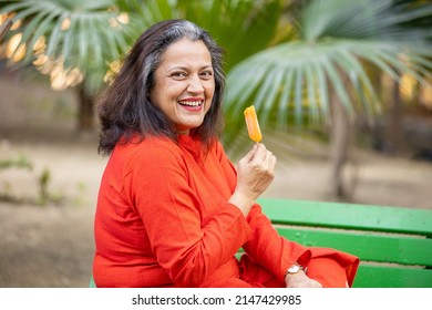 Portrait of happy indian senior woman eating ice lolly or ice cream in a park outdoor, old mature lady enjoy retirement life. summer holidays.