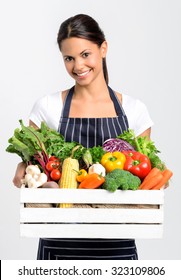 Portrait of happy indian woman chef holding a crate full of fresh organic vegetables on grey background, promoting eating seasonally and sourcing from local producers