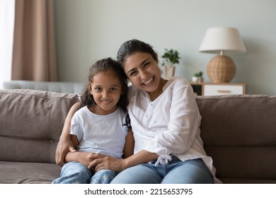 Portrait Of Happy Indian Woman Hugs Little Adorable Daughter Laughing Seated Together On Sofa Staring At Camera Enjoy Pleasant Communication Through Video Conference App. Family Bond, Motherhood, Fun