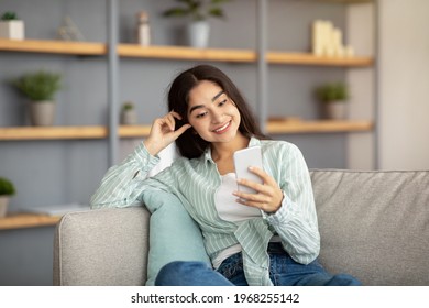 Portrait Of Happy Indian Woman Using Smartphone On Couch In Living Room, Checking Her Email, Posting In Social Media. Young Lady With Mobile Device Browsing Web, Watching Video Or Communicating Online