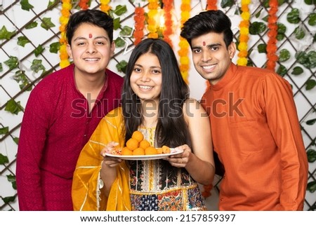 Portrait of happy indian young kids or bothers and sister wearing traditional cloths holding plate full of laddu or laddoo sweets celebrating diwali festival looking at camera with smile.