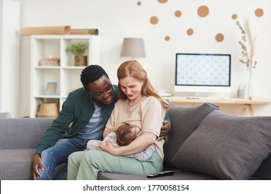 Portrait of happy interracial family sitting on couch at home together with mature mother breastfeeding baby, copy space