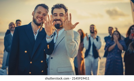 Portrait Of A Happy Just Married Handsome Gay Couple Kissing, Showing Off Their Gold Wedding Rings. Two Attractive Queer Men In Suits Smile And Pose For Camera With Friends. LGBTQ Family Goals.