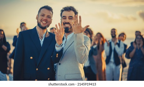 Portrait Of A Happy Just Married Handsome Gay Couple Kissing, Showing Off Their Gold Wedding Rings. Two Attractive Queer Men In Suits Smile And Pose For Camera With Friends. LGBTQ Family Goals.