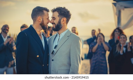 Portrait Of A Happy Just Married Handsome Gay Couple Kissing, Showing Off Their Gold Wedding Rings. Two Attractive Queer Men In Suits Smile And Pose For Camera With Friends. LGBTQ Family Goals.