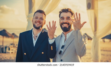 Portrait Of A Happy Just Married Handsome Gay Couple In Love Showing Off Their Gold Wedding Rings. Two Attractive Queer Men In Suits Smile And Pose For Camera. LGBTQ Relationship And Family Goals.