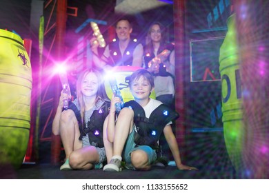 Portrait Of Happy Kids Sitting In Bright Beams With Laser Guns During Laser Tag Game With Parents
