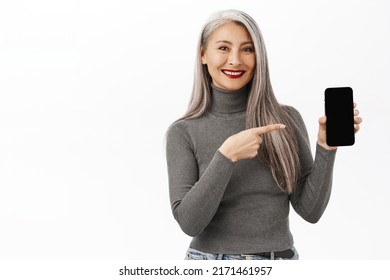 Portrait Of Happy Korean Senior Woman, Beautiful Middle Aged Female Model Pointing At Smartphone App, Showing Mobile Screen, Standing Over White Background