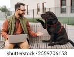 Portrait of happy Latin man wearing eyeglasses, casual clothes, using laptop, playing with black labrador, holding his paw. Animals, love pet concept