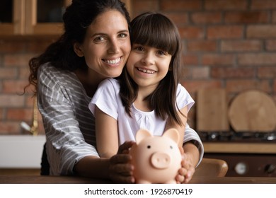 Portrait Of Happy Latino Young Mom And Small 8s Daughter Recommend Smart Living And Saving. Smiling Hispanic Mother And Little Teen Girl Child Hold Piggybank Feel Economical Making Investment.