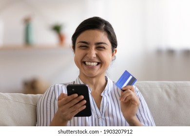 Portrait of happy laughing indian female looking at camera holding phone plastic card enjoy buying shopping process. Young lady having pleasure in spending money online prepaying internet store orders