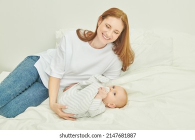 Portrait Of A Happy, Laughing Mother Lying On A Bed With A Cute Baby. Happy Motherhood. Mother And Child.