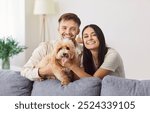 Portrait of happy laughing young couple close near couch with cute little dog, smiling husband and wife with puppy, relaxing together in new own living room, apartment renters with companion animal 